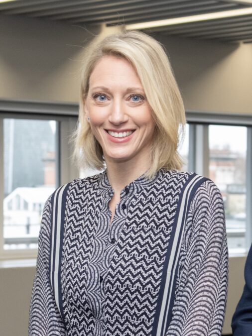 A photograph of Claire Waxman smiling at the camera. She is wearing a patterned blue and white collared shirt. The background is a blurred office setting with windows.